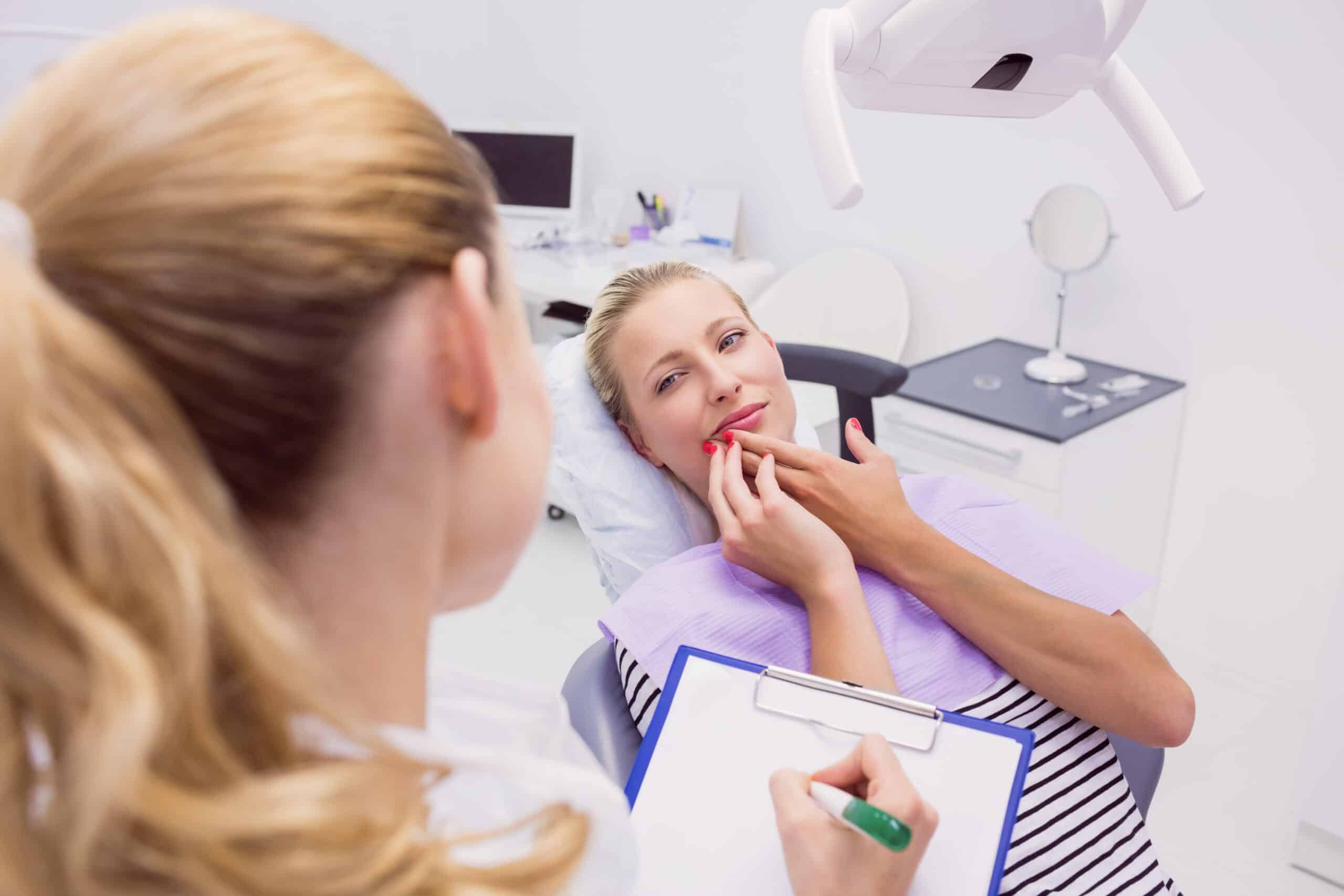 Female patient in dental office discussing toothache with dentist, holding cheek in discomfort, clipboard and dental equipment visible.