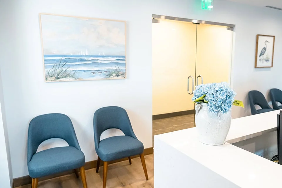 Waiting area of West Periodontics and Dental Implants featuring blue chairs, a vase of hydrangeas, and a coastal painting on the wall.
