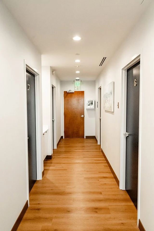Modern dental office hallway with wooden flooring, featuring multiple doors labeled with room numbers, leading to treatment areas at West Periodontics and Dental Implants.